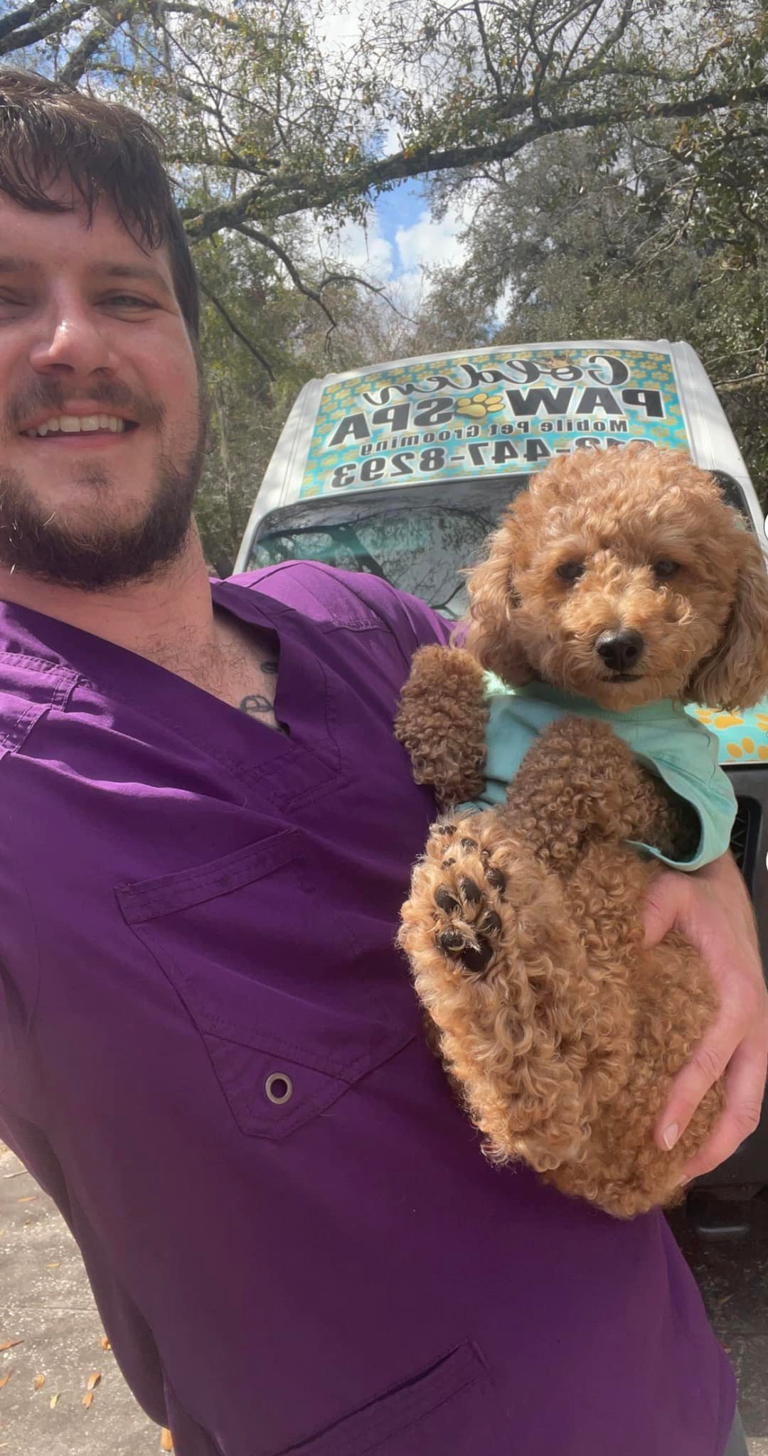 Groomer in purple scrubs holding a small dog in front of the Golden Paw Spa van